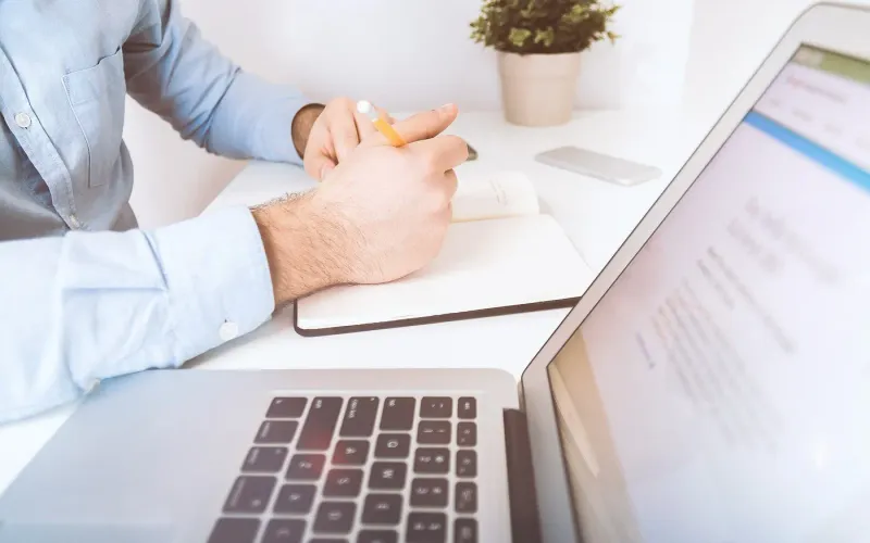 Person writing notes in a notebook next to an open laptop